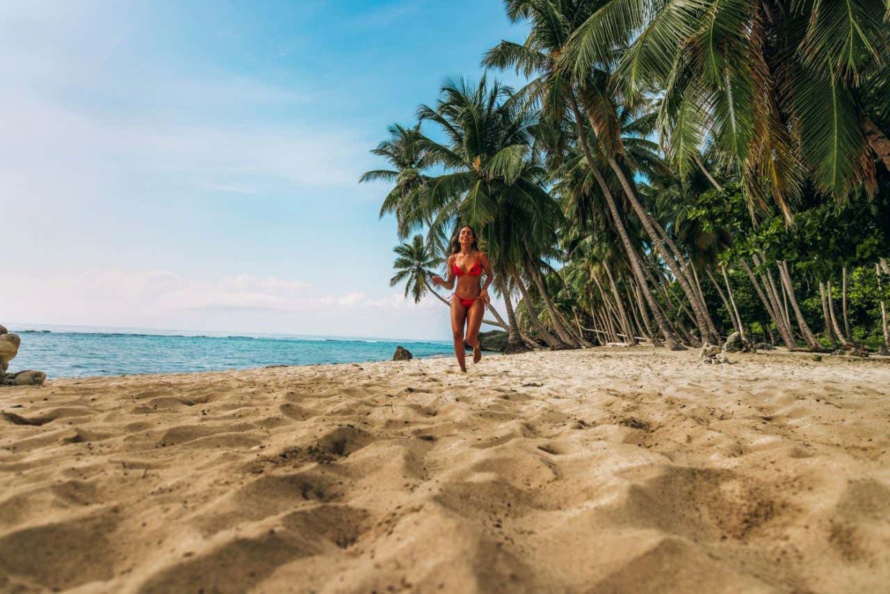Una mujer corre en una playa de arena de tonos marrón con unas palmeras al fondo