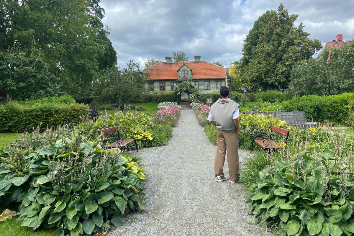 Un hombre de espaldas a la cámara contemplando una casa de madera de color verde en un jardín con muchas plantas y flores
