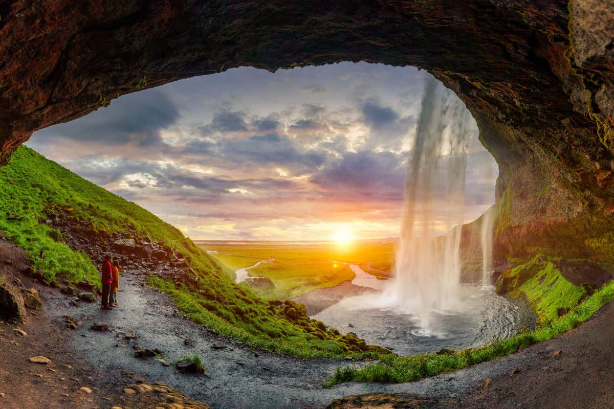Una pareja en mitad de una cueva contemplando el atardecer y viendo cómo cae el agua por una cascada de Islandia