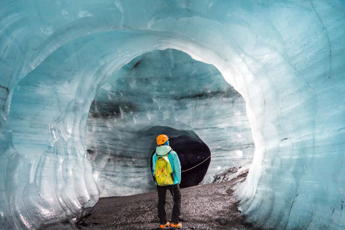 Un hombre de espaldas a la cámara con equipamiento de trekking contemplando el interior de una cueva cuyas paredes son todas de hielo