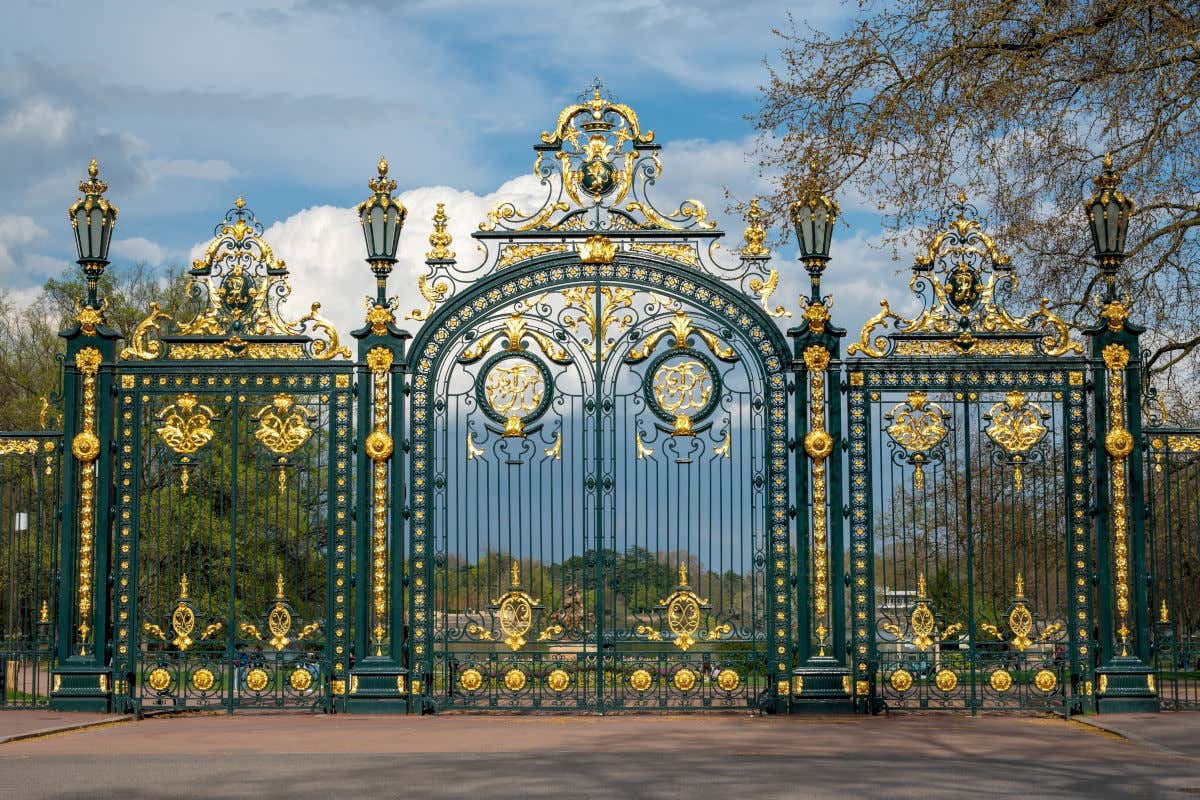 Un pórtico de hierro decorado con tonos verdes y dorados que precede a un parque con varios árboles y un estanque