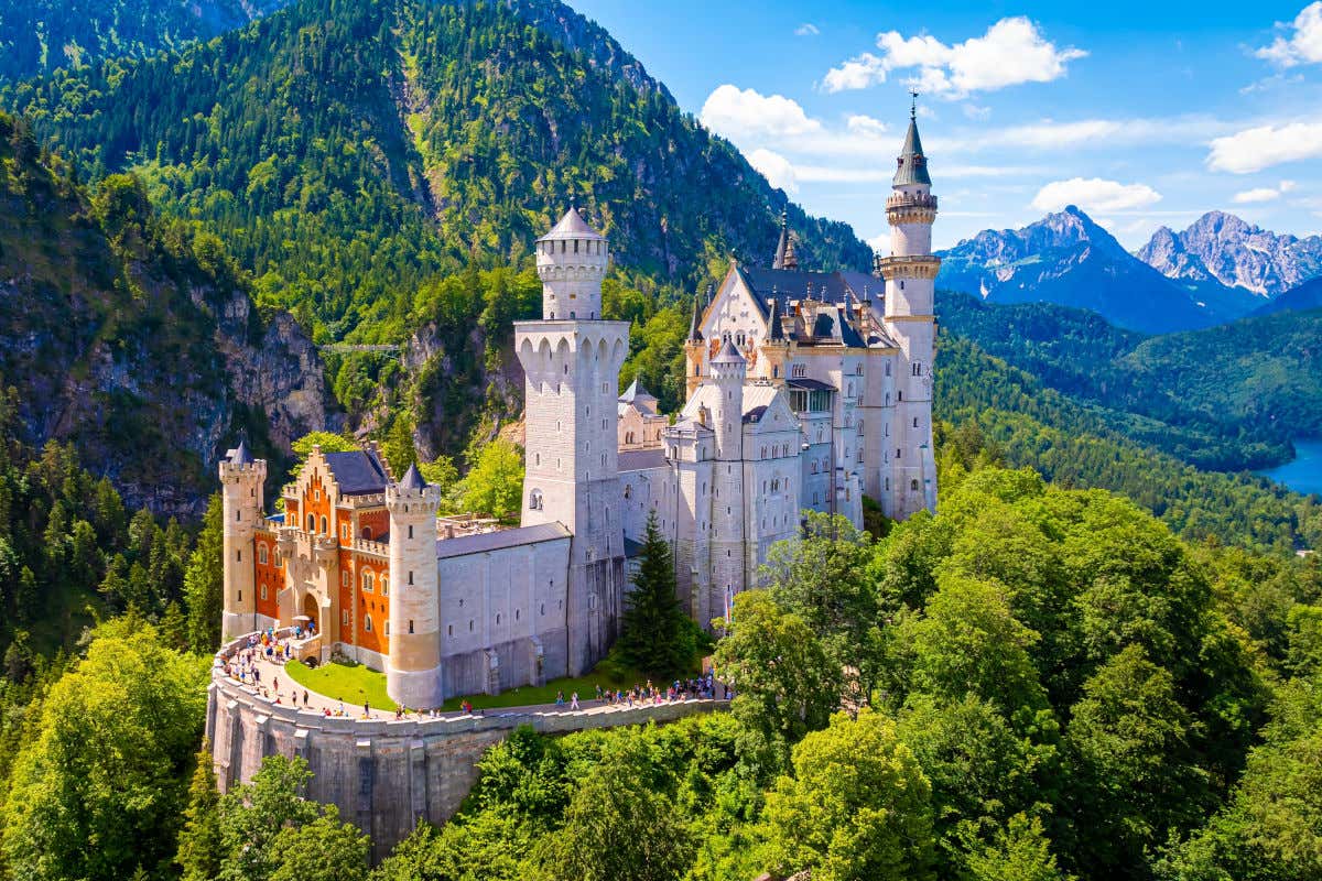 El castillo de Neuschwanstein con numerosas torres y torreones en mitad de una colina rodeada de árboles, montañas y un lago