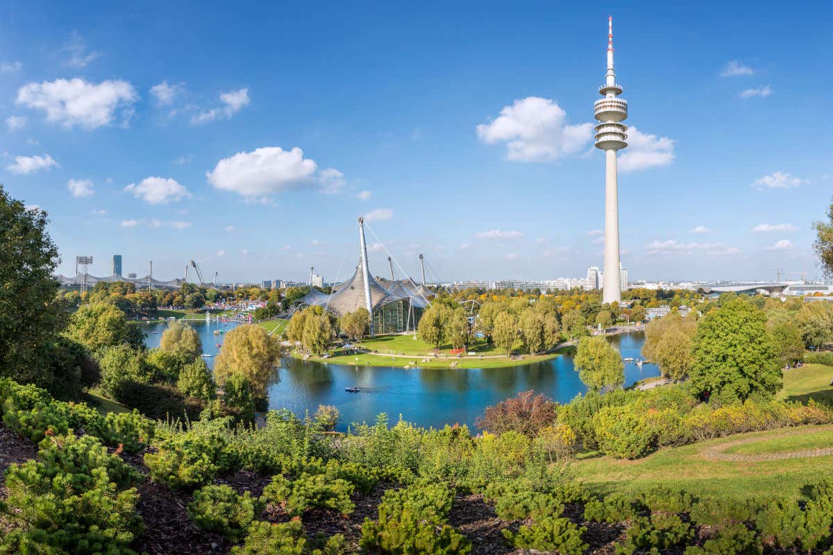 Panorámica del parque olímpico de Múnich repleto de árboles, con un río de aguas cristalinas y con varias estructuras de recintos deportivos juntos a una gran torre de televisión