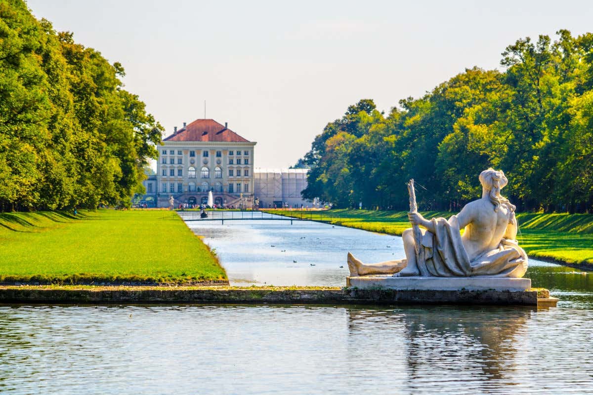 El palacio de Nymphenburg frente a un estanque rodeado de árboles verdes y con una estatua femenina decorando la escena