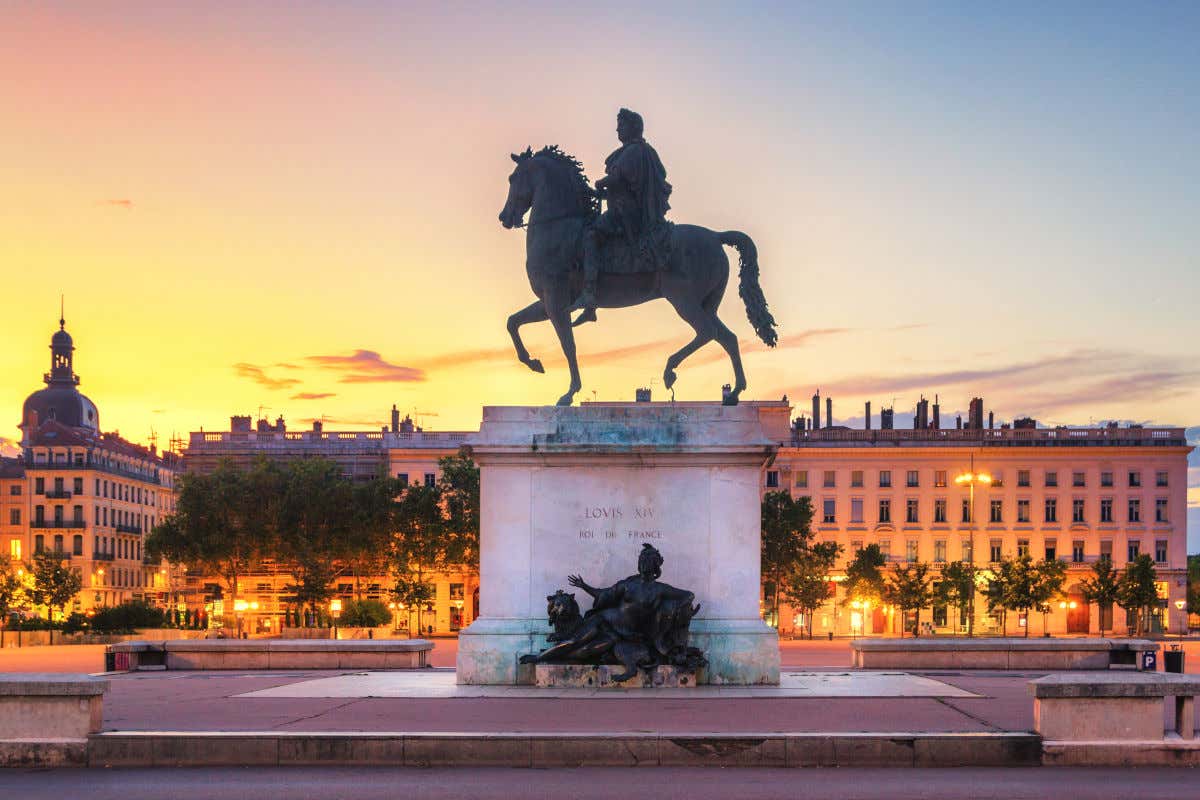 Escultura ecuestre de bronce del rey Luis XIV en mitad de una plaza rodeada de edificios que se van iluminando al atardecer