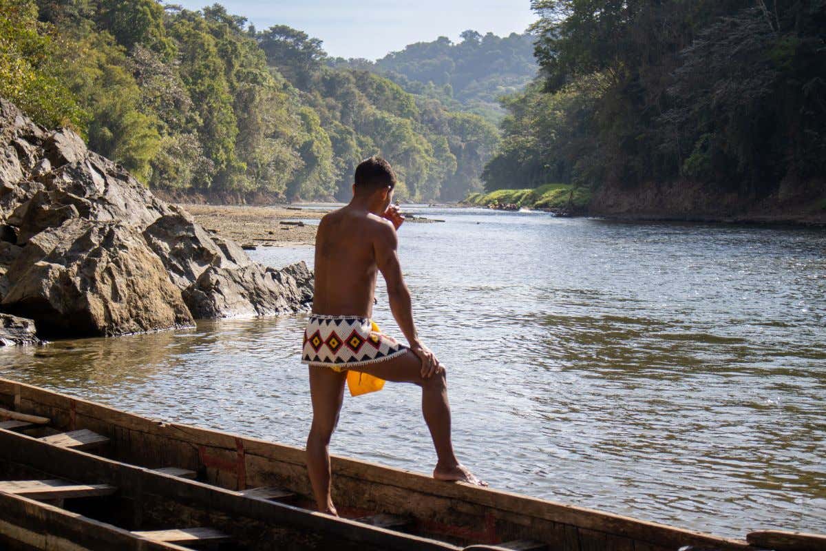 Un hombre de espalda sobre una lancha de madera en indumentaria indígena viendo un río