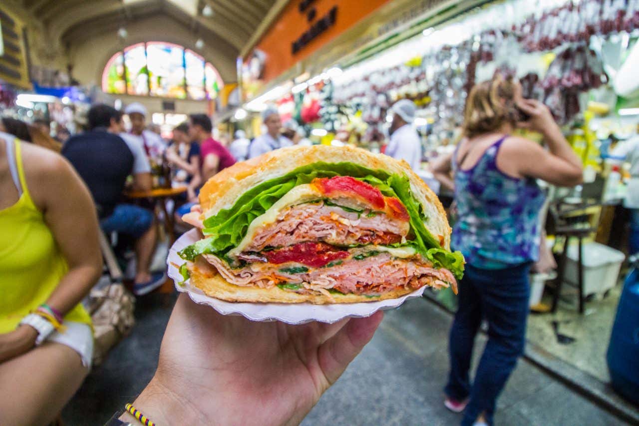 A hand holds up a paper plate with a stuffed mortadella sandwich on it, lettuce, cheese tomatoes visible in cross section, bustling market stalls behind