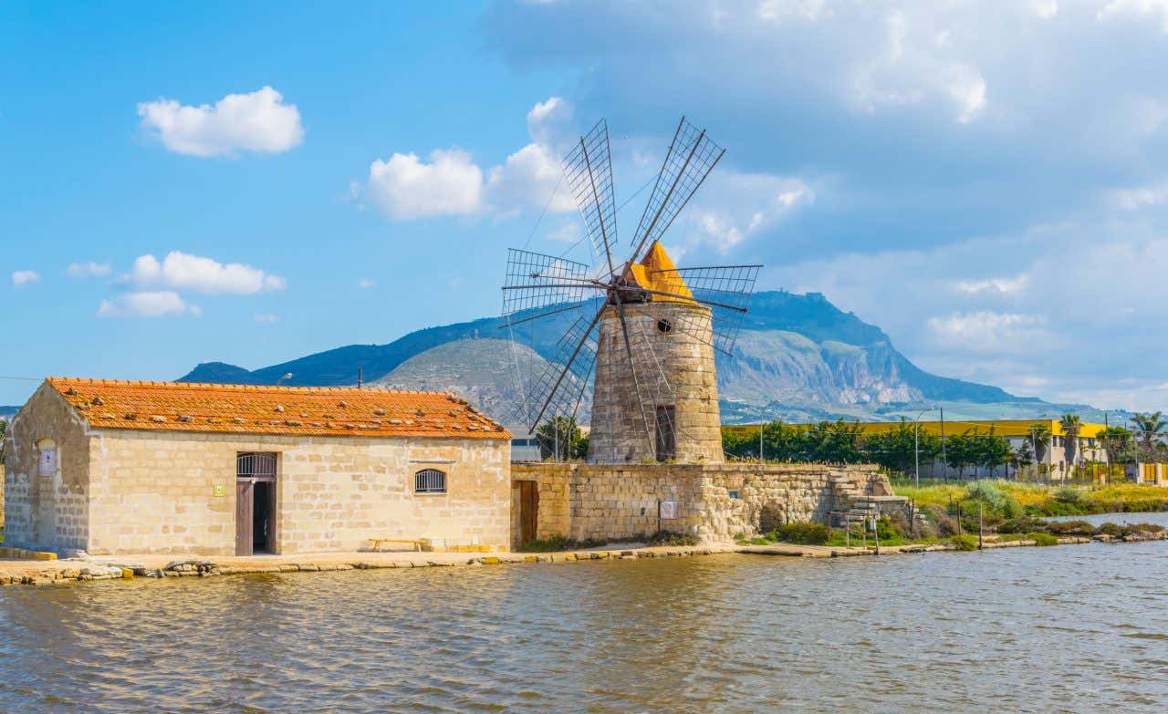 Un mulino a vento in pietra accanto a una bassa costruzione in mattoni nei pressi di una salina di Trapani, sullo sfondo un alta montagna verde in una giornata parzialmente nuvolosa
