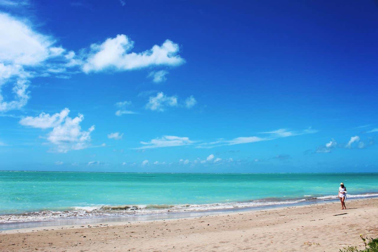 Uma mulher caminhando em uma bela praia com águas cristalinas e verdes em um dia ensolarado com céu azul, um excelente plano do que fazer em Porto de Galinhas