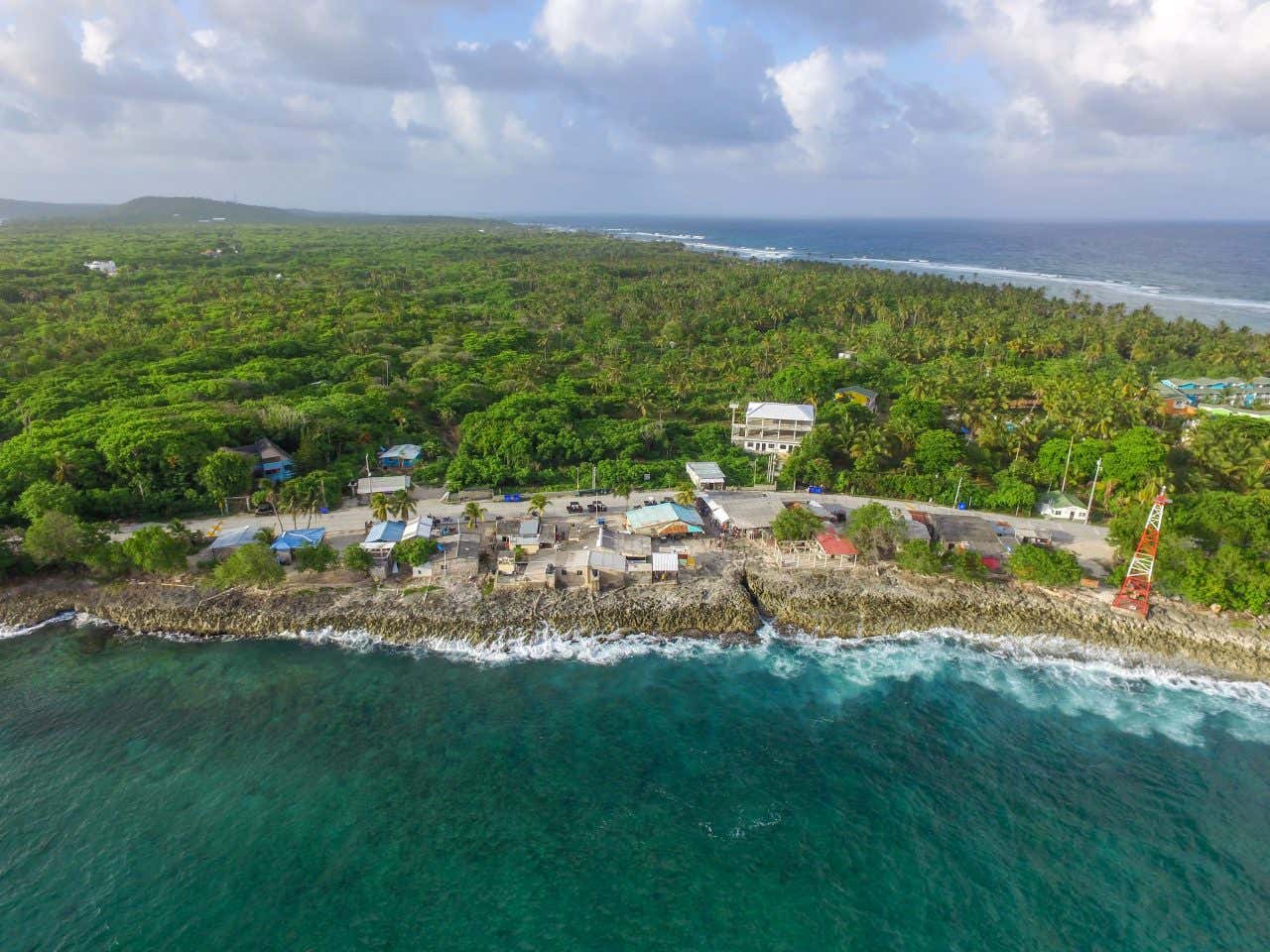 An aerial view of a coastal town or village situated on a rocky shoreline. Waves are breaking on the rocks, and a road with a few small buildings runs parallel to the coast. Dense green foliage covers the area behind the town.