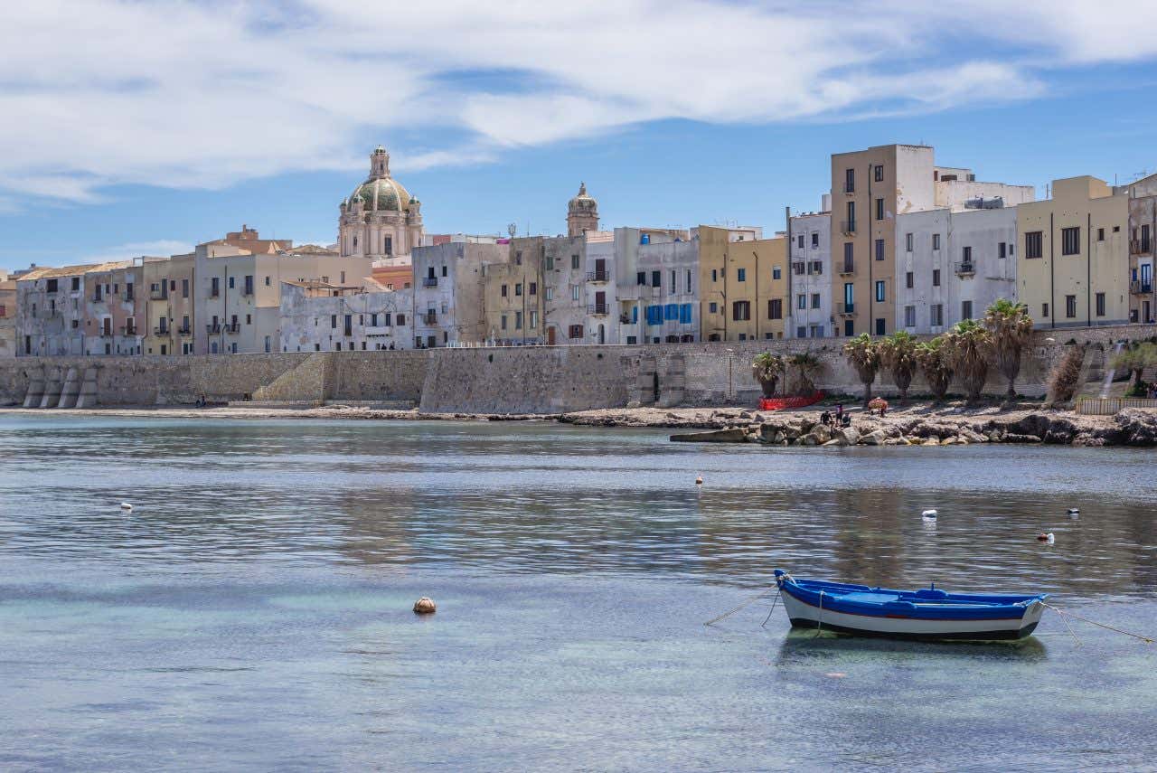Scorcio su Trapani vista dal mare, con l'acqua placida e una piccola barchetta di legno ancorata nei pressi di una caletta rocciosa, sullo sfondo le passe mura di cinta medievali dividono i palazzi dal mare