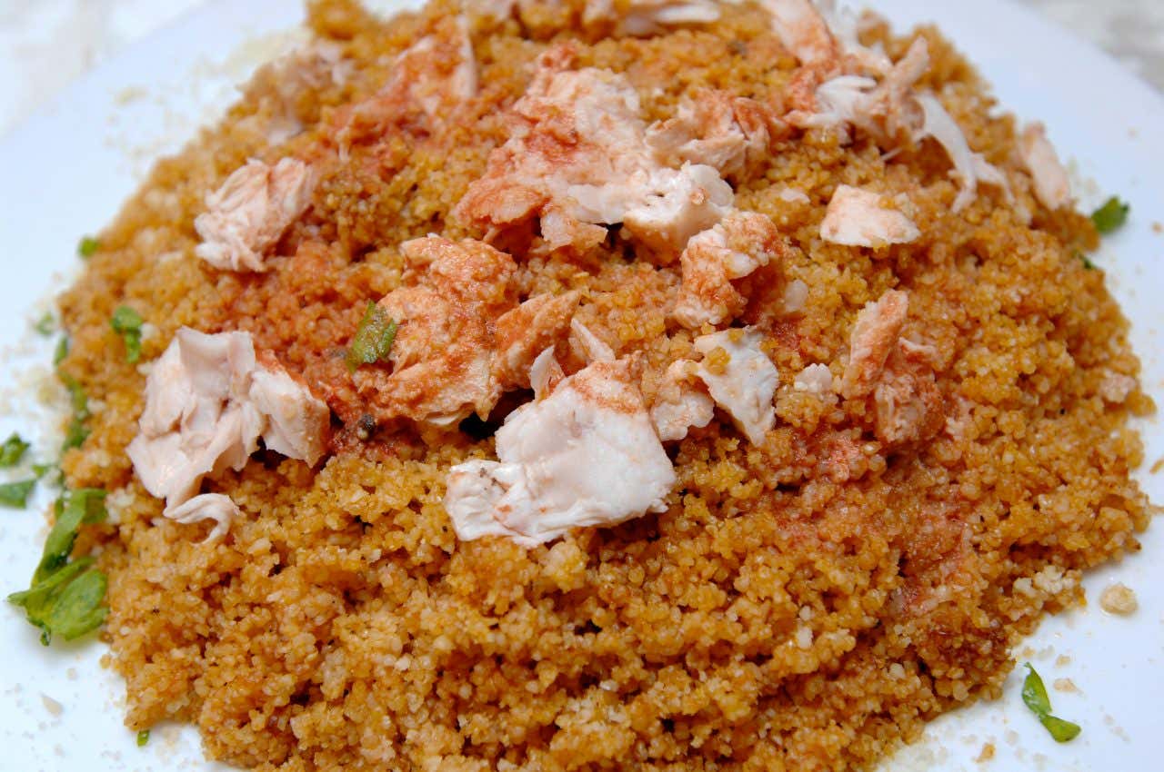 Un piatto di cous cous dal colore rosso, condito con pesce fresco bollito e prezzemolo