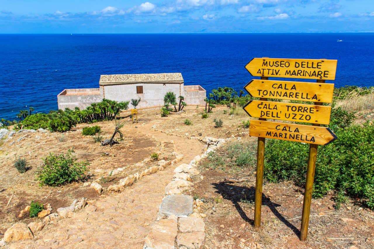 Vista sul mare da una collina brulla nella Riserva dello Zingaro. In primo piano ci sono quattro cartelli di legno che segnalano la direzione per le calette della riserva, mentre sullo sfondo un sentieri di terra conduce a una piccola costruzione in mattoni, mentre all'orizzonte si staglia il mare blu