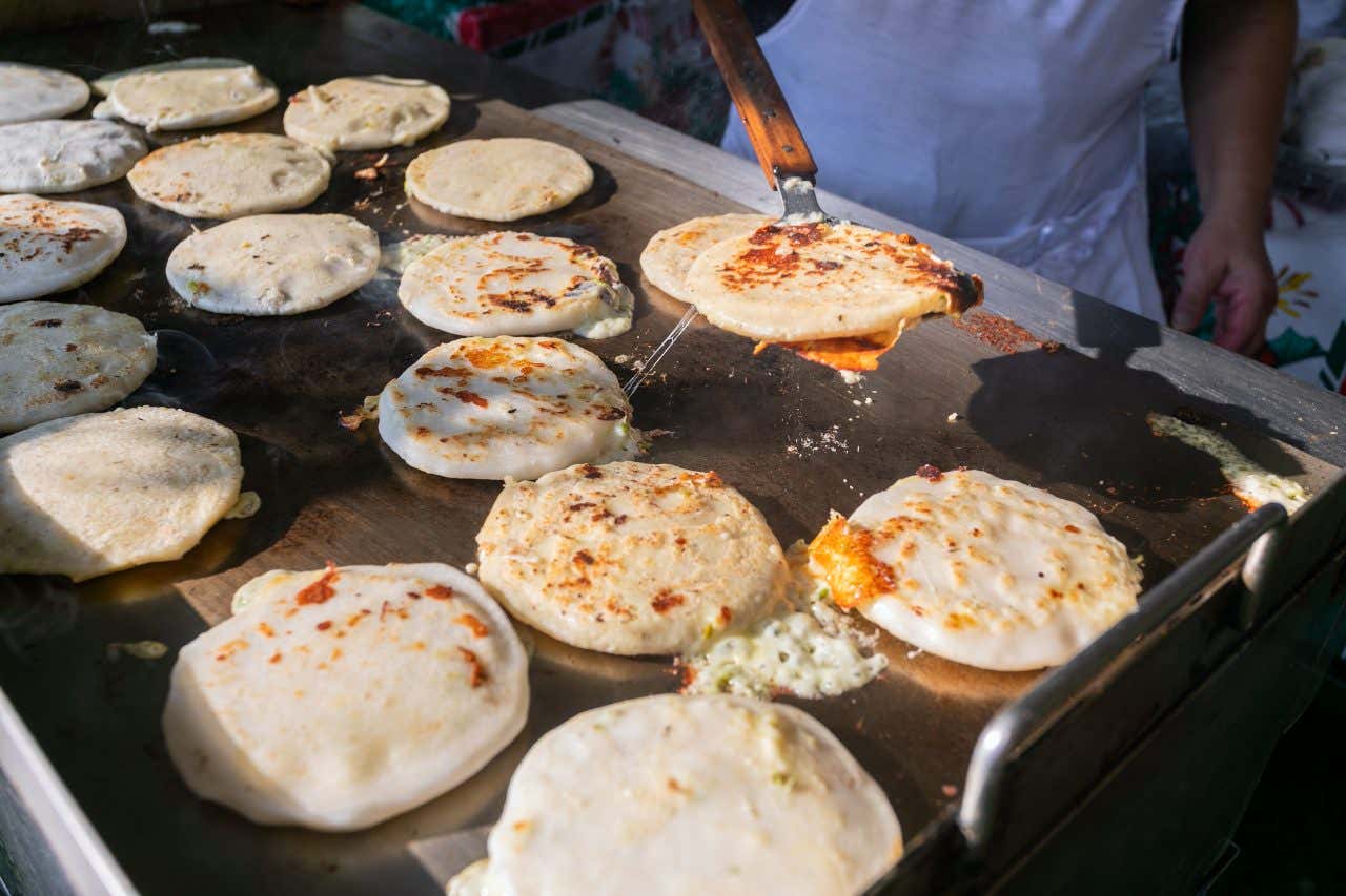 A grill filled with freshly made pupusas, a cook visible in the background flipping the shells over in San Salvador, El Salvador, a secret foodie destination