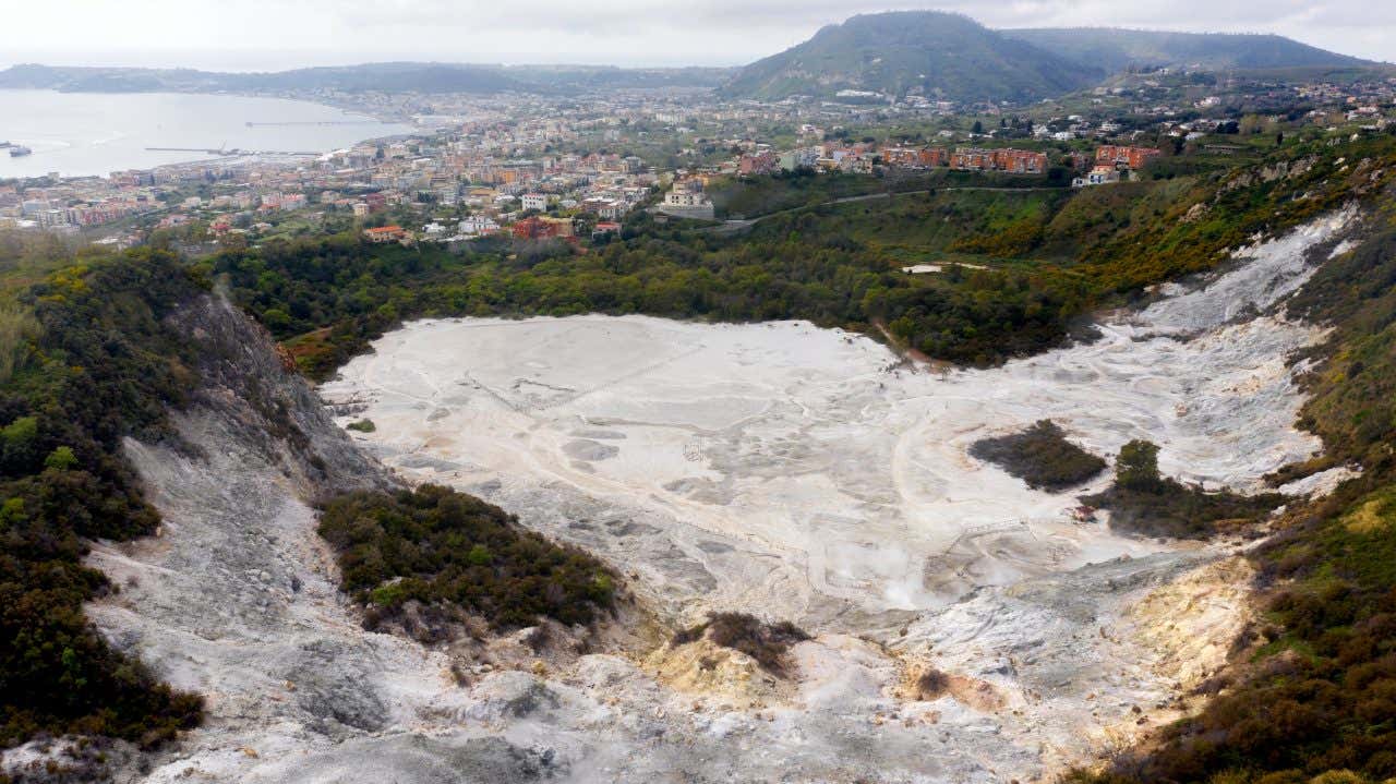 Veduta panoramica della Solfatara di Pozzuoli, un cratere vulcanico fatto di terra bianca sulfurea fumante, circondato da vegetazione, con la città di Pozzuoli e il mare sullo sfondo