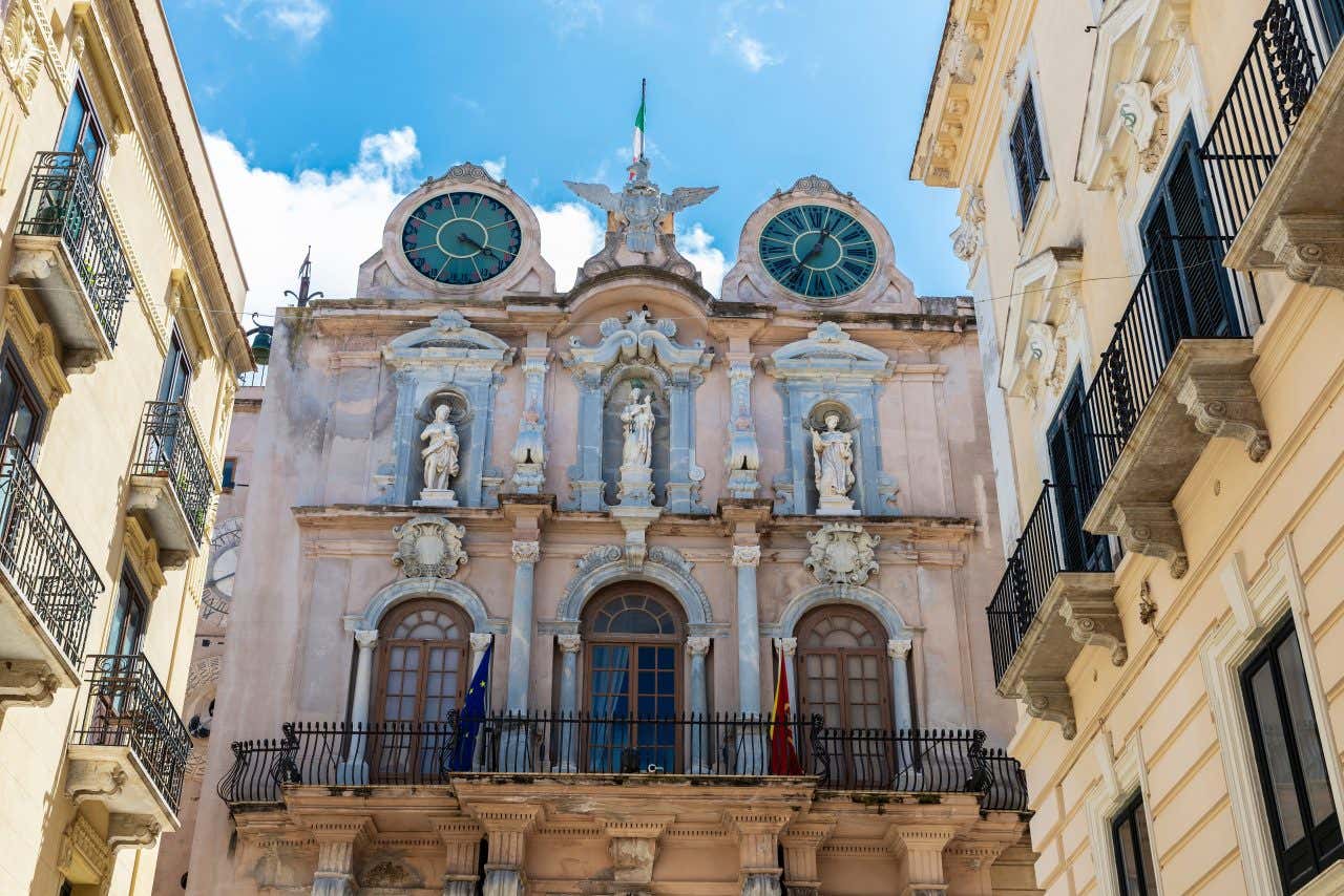 Facciata di un palazzo in stile barocco nel centro storico di Trapani fotografato dal basso. La facciata è divisa in due piani, decorati con statue e colonne, mentre la cima dell'edificio ospita due orologi
