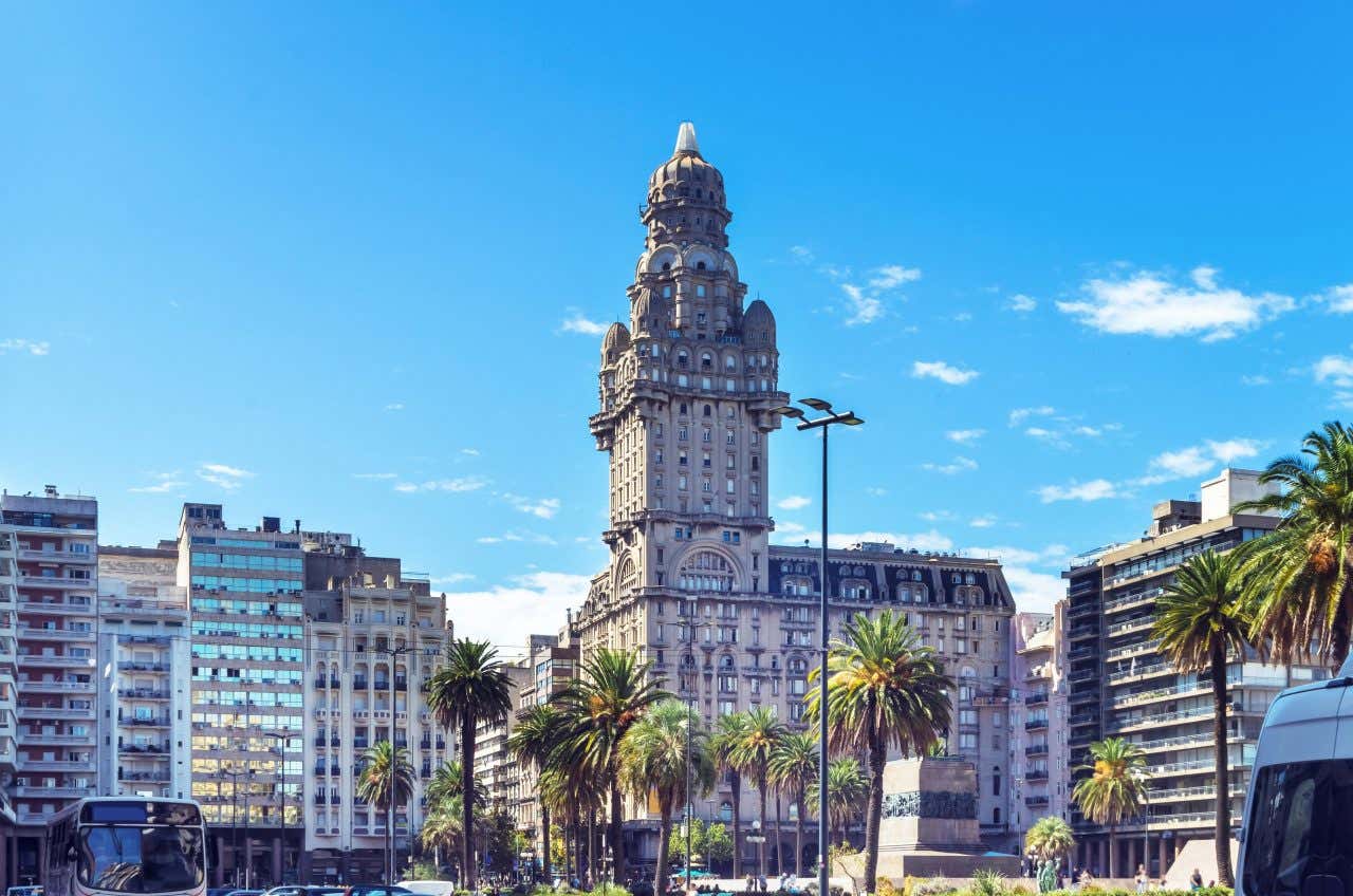 The cityscape of Montevideo seen from street level with palm trees on the street and a cloudy blue sky in the background.