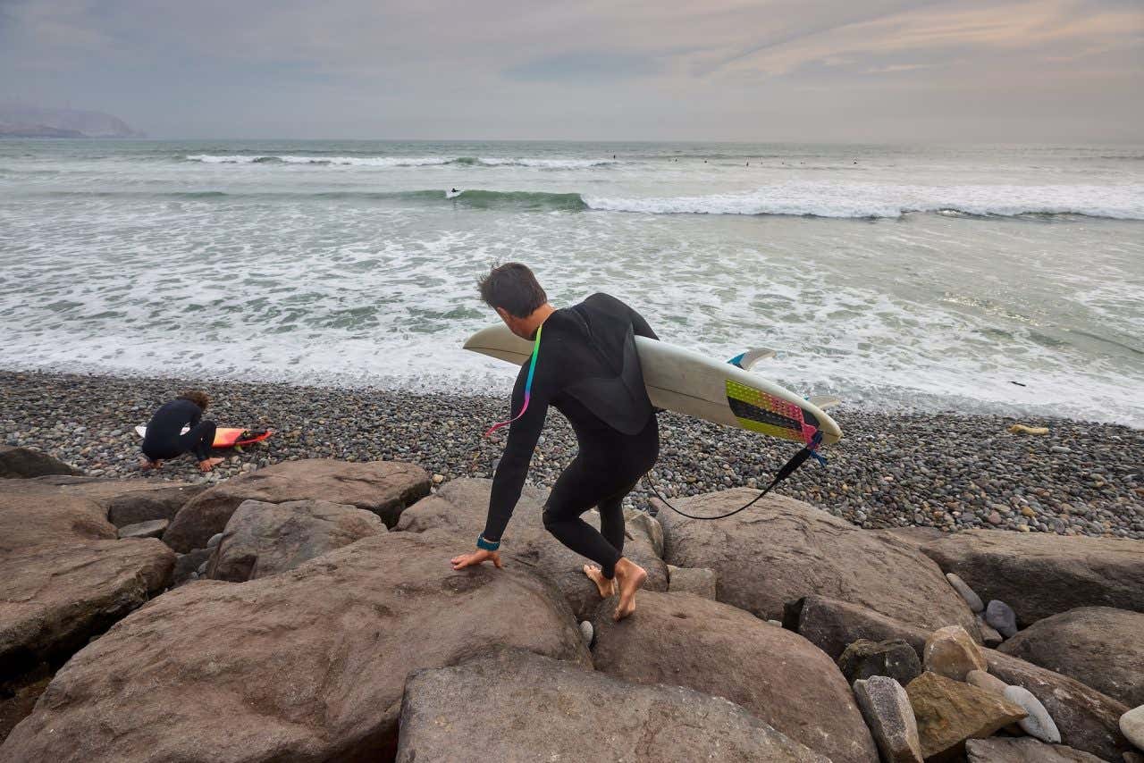 Dois homens com pranchas descendo pela pedra e se preparando para entrar ao mar