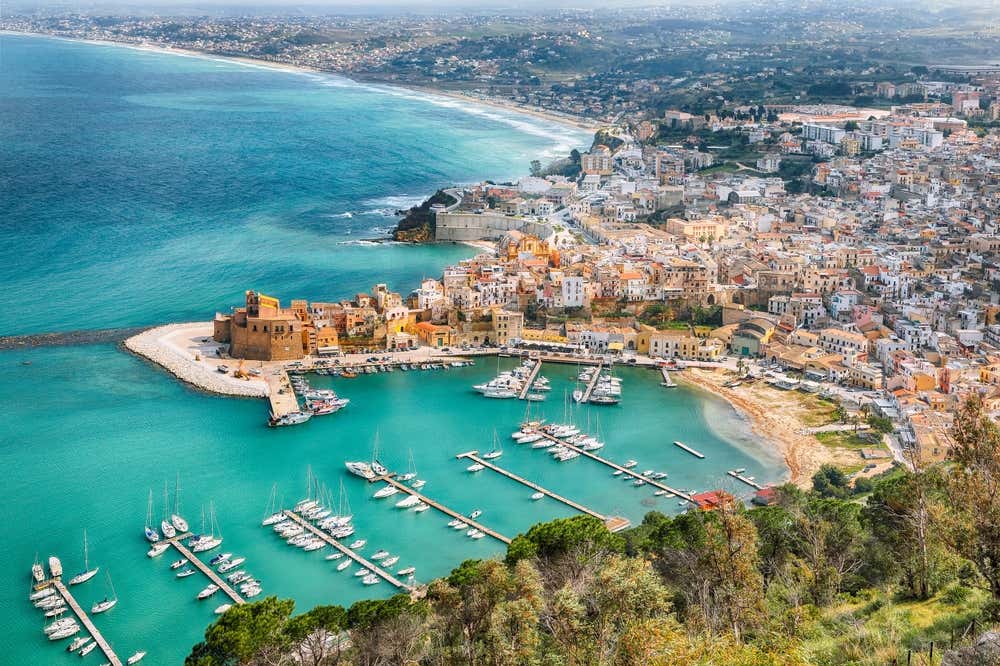 Veduta aerea panoramica di Trapani, Sicilia, che mostra il porto turistico con numerose barche a vela ormeggiate nelle acque turchesi cristalline, il centro storico con edifici color ocra e pastello, tra cui fortificazioni storiche in primo piano, e la lunga spiaggia che si estende lungo la costa con la città che si sviluppa verso l'entroterra