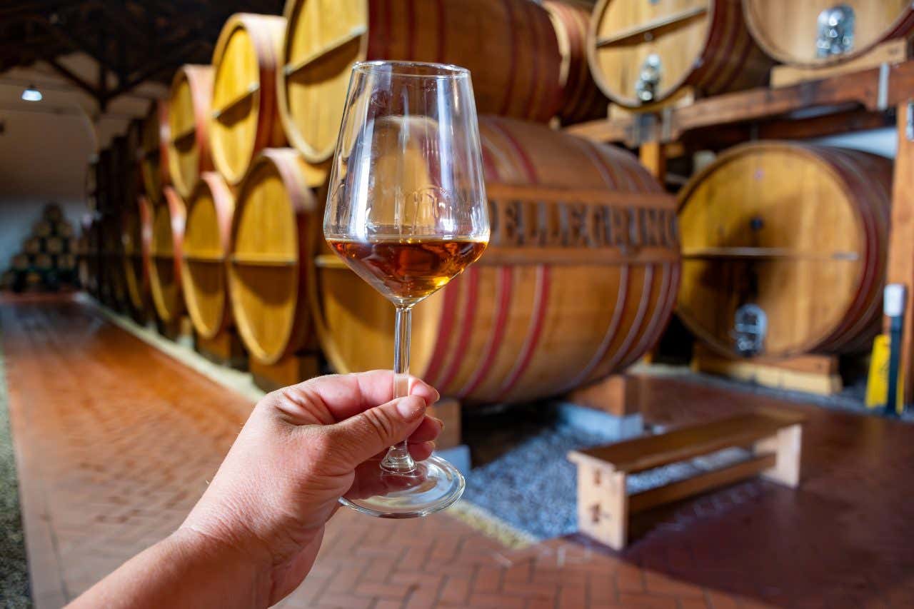 Una mano in primo piano regge un calice di vino Marsala dal colore ambrato, all'interno di una cantina con botti di legno