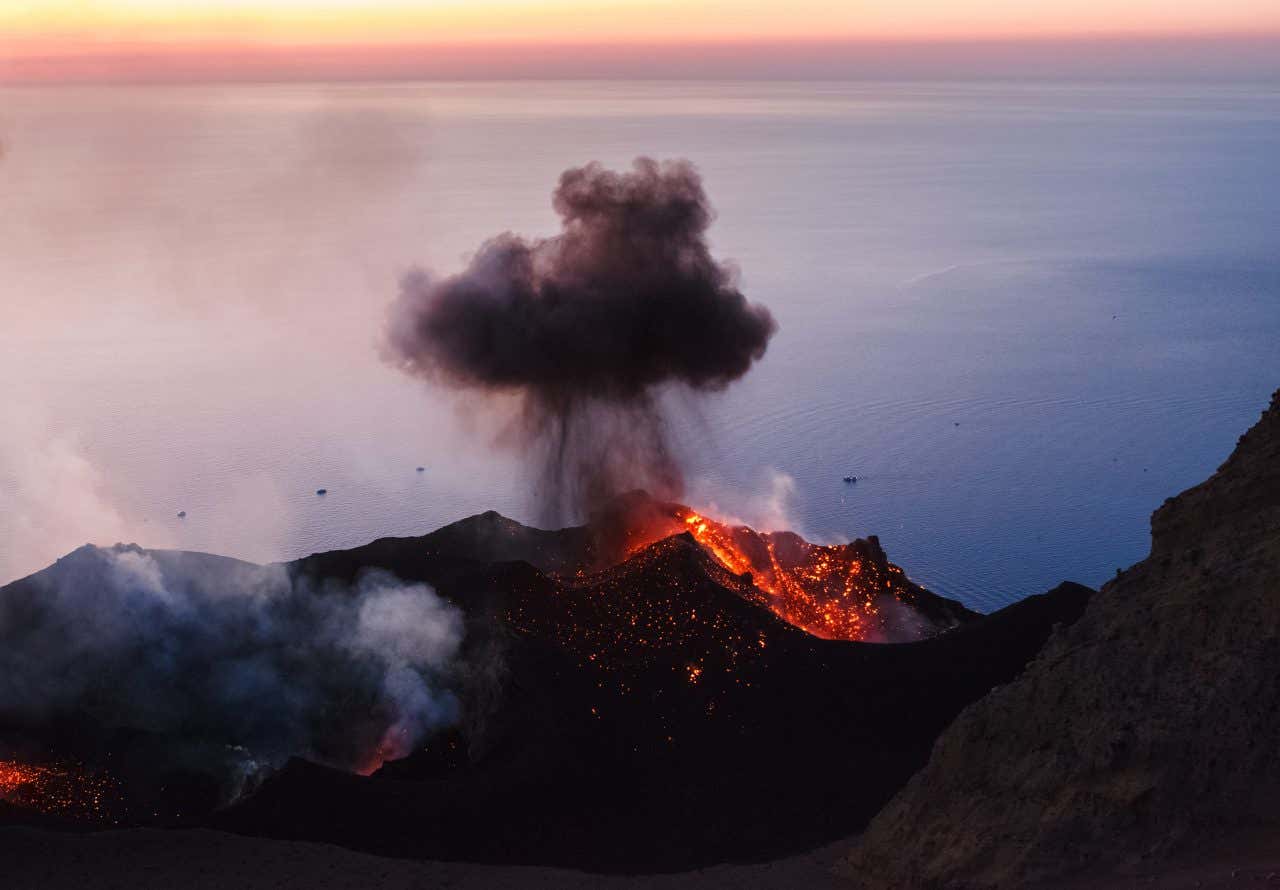 Foto ravvicinata del cratere del vulcano Stromboli durante un'eruzione: uno sbuffo di fumo nero risale dalla bocca del cratere pieno di lava incandescente, sullo sfondo il mare al tramonto con alcune imbarcazioni