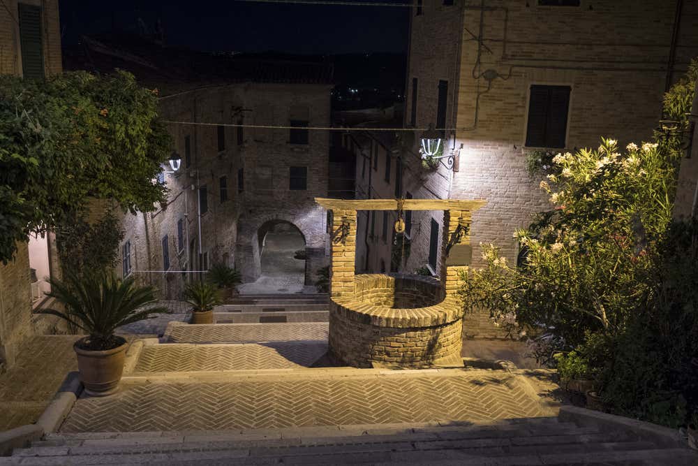 Il Pozzo della Polenta, in mattoni, lungo la scalinata della Piaggia a Corinaldo, di notte. Intorno ci sono gradini con pavimentazione a spina di pesce, case in mattoni illuminate dai lampioni, un arco in fondo al vicolo e piante di oleandro in fiore
