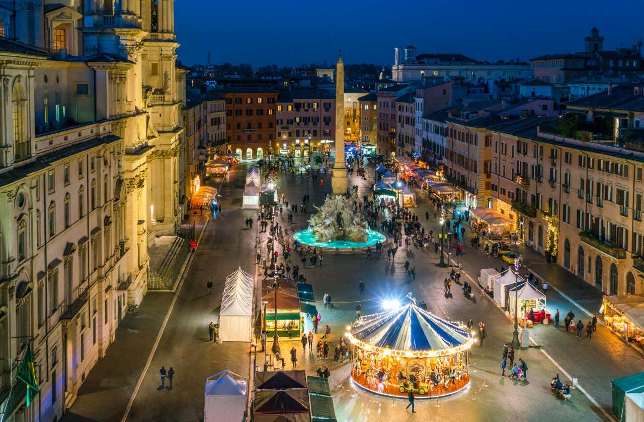 Piazza Navona ripresa dall'alto di sera, con al centro della piazza, stand di mercatini e una giostra. Le persone passeggiano e osservano la merce esposta