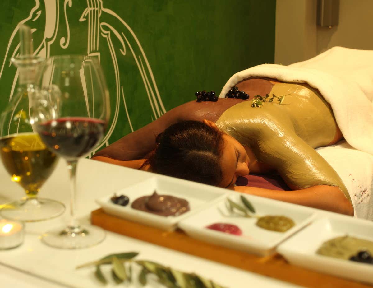 Mujer tumbada con la espalda cubierta por una mascarilla de barro y unas copas de vino en primer plano