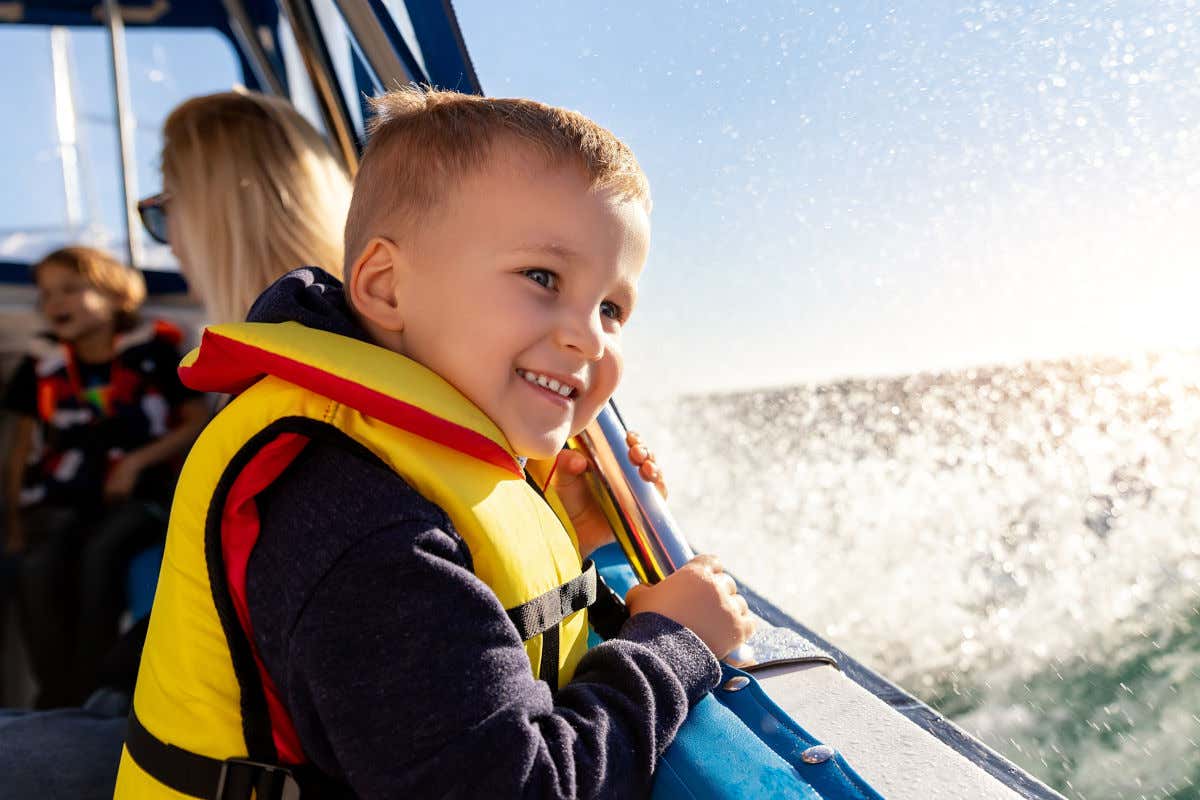 Un niño con chaleco salvavidas subido en un barco y contemplando alegremente cómo se mueven las olas