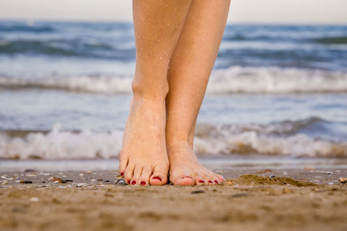 Unos pies femeninos con las uñas pintadas pisando la arena dorada de una playa con las olas del mar rompiendo muy cerca