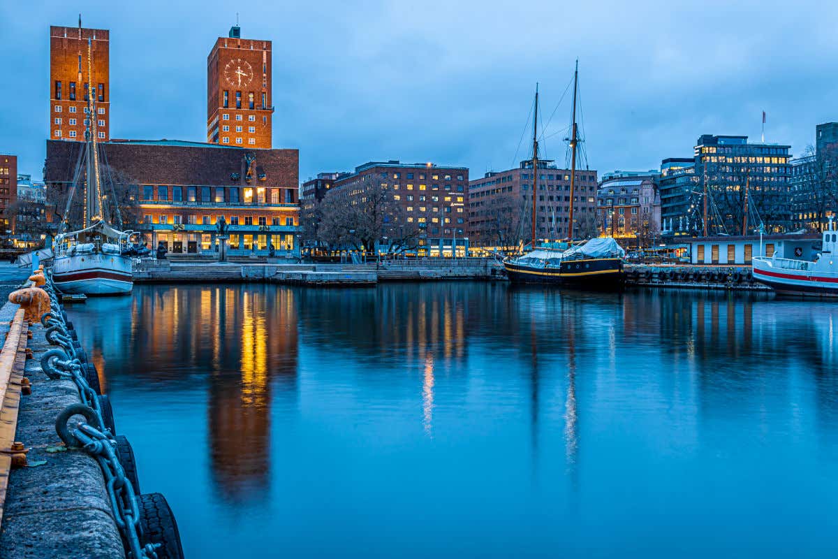 El puerto de Oslo con el mar en calma y varios barcos frente al Ayuntamiento de la ciudad, un edificio de ladrillo rojo con dos altas torres