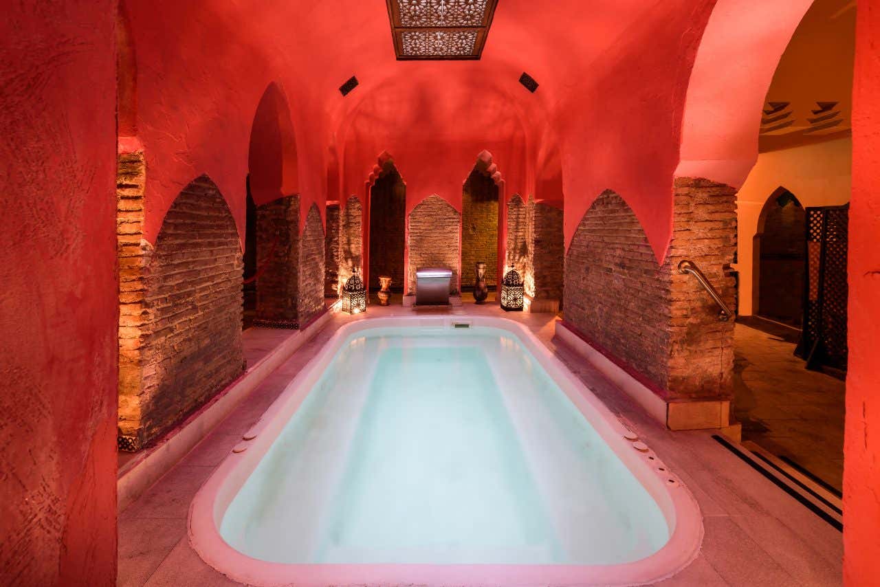  Interno di un hammam tradizionale di Granada con vasca termale centrale illuminata da luce blu, archi moresco-andalusi in mattoni rossi, soffitto a volta rosa corallo, lanterne decorative in ferro battuto e atmosfera calda e rilassante tipica dei bagni arabi storici della città