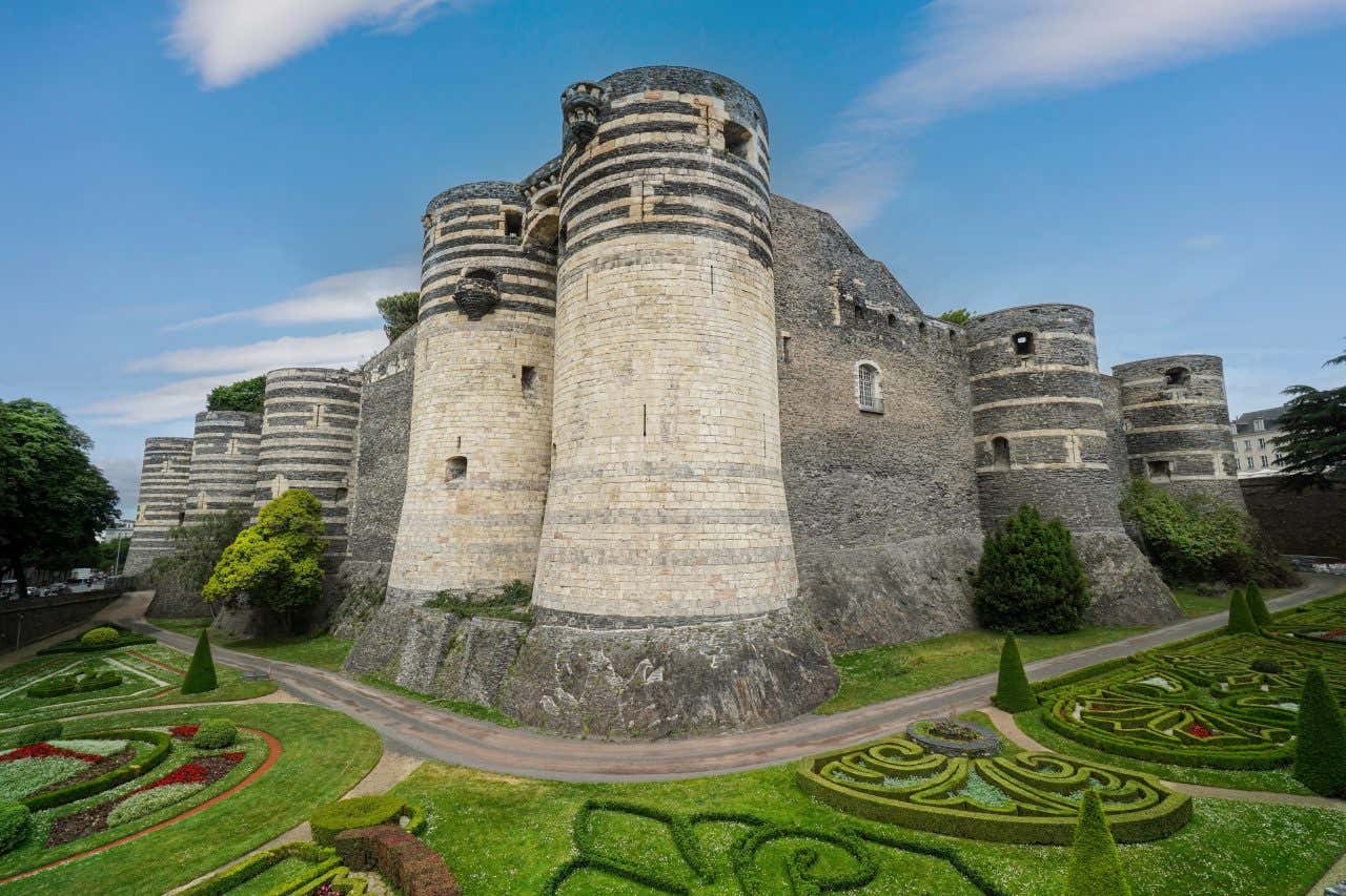 The Château d'Angers, a large medieval fortress with multiple round towers made of grey and white striped stone, is surrounded by manicured gardens with hedges and floral patterns. The castle stands under a blue sky with some clouds.
