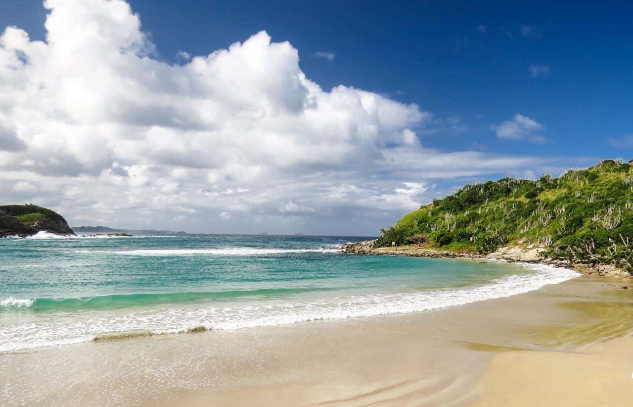 Paisagem de praia com areia dourada, águas turquesa e montes cobertos com vegetação ao redor