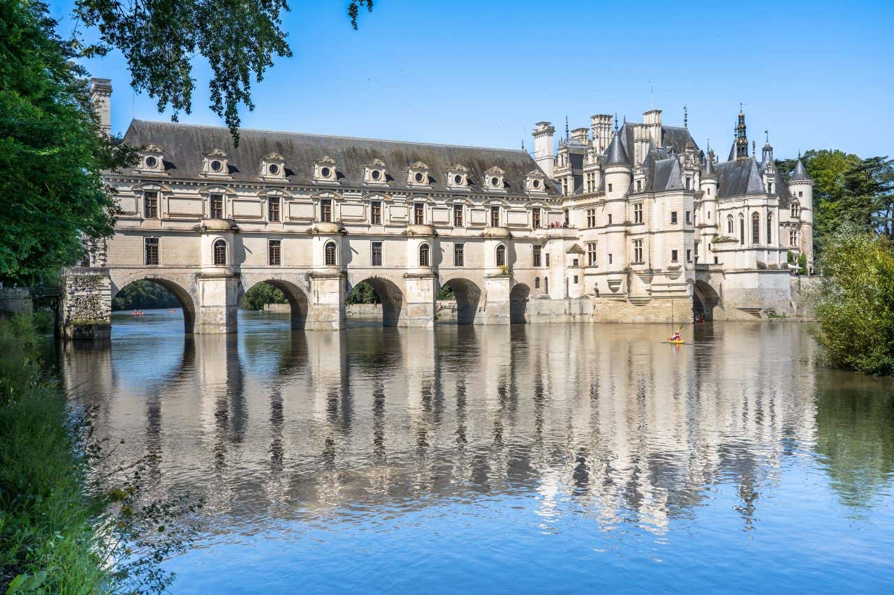 The Château de Chenonceau, a French castle that spans a river with its arched bridge-like structure. The white stone building reflects in the calm water on a sunny day.