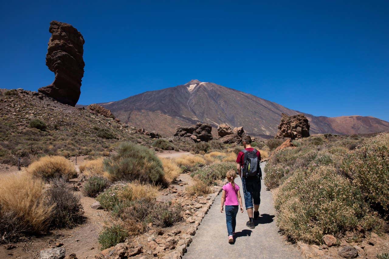 A father and a daughter walking in Teide National Park on a sunny day, with cliffs and Mount Teide in the distance.