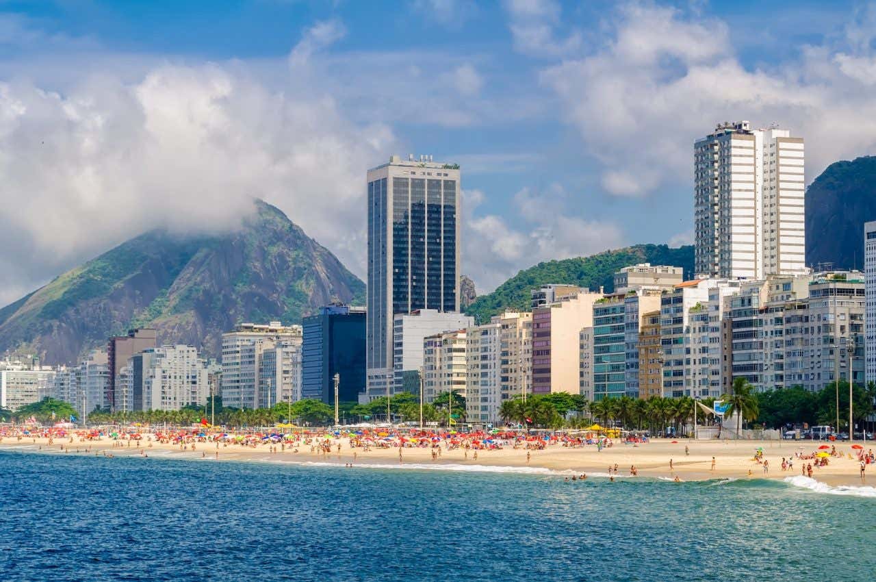 Vista do mar à Copacabana, com mutos banhistas, guarda-sóis, prédios e monstes ao fundo