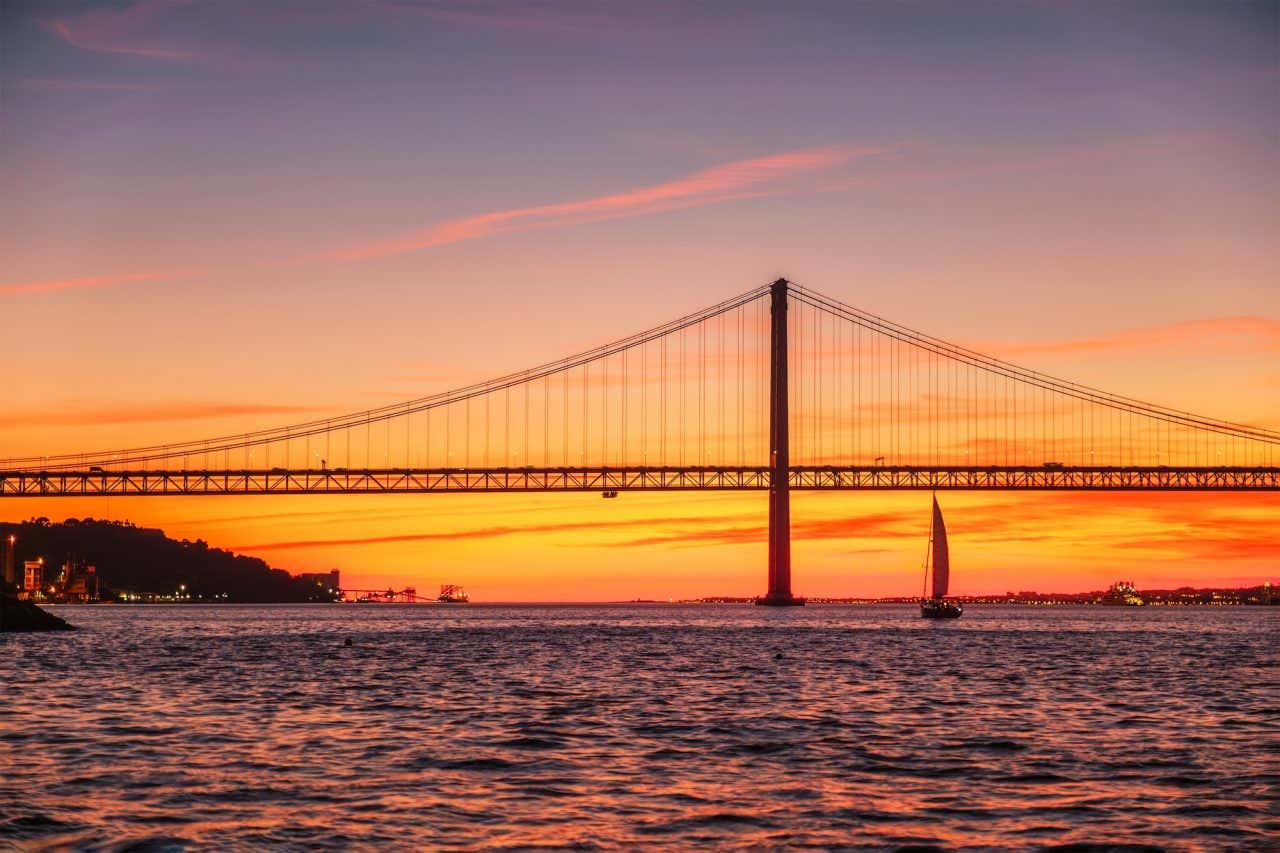 Vista da Ponte 25 de Abril, famoso marco turístico de Lisboa, que liga Lisboa e Almada sobre o rio Tejo, com a silhueta de um iate turístico ao pôr do sol