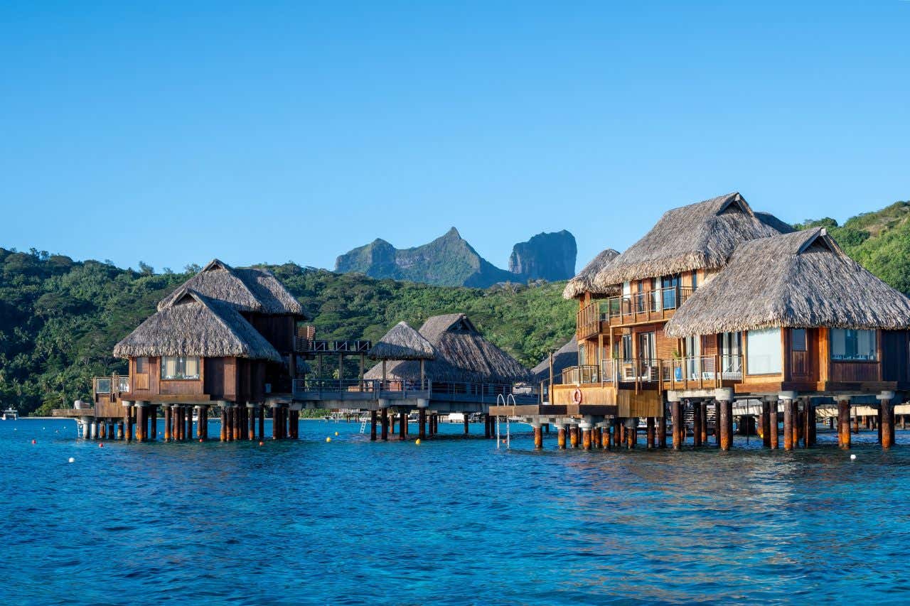 Un gruppo di case in legno con un tetto di paglia costruite sopra palafitte sull'isola di Bora Bora, con la cima di una montagna vulcanica sullo sfondo