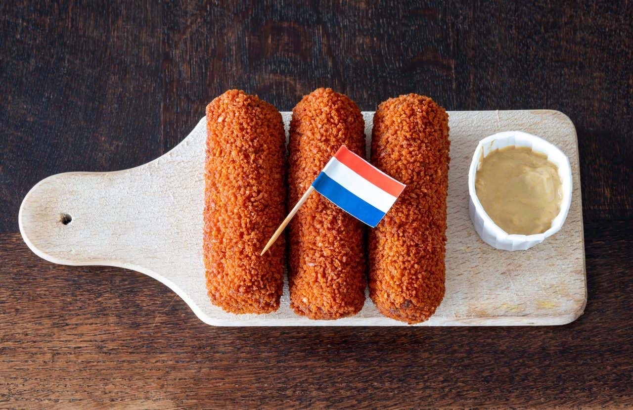 Três croquetes sobre uma tábua de madeira e uma mini bandeira holandesa em cima acompanhados de mostarda
