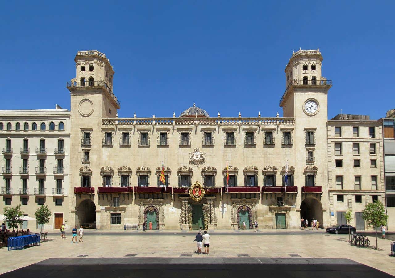 Facciata del municipio di Alicante, in Spagna, in stile barocco, con due torri laterali, orologio sulla torre di destra, balconate con bandiere ufficiali e una grande piazza antistante sotto un cielo azzurro