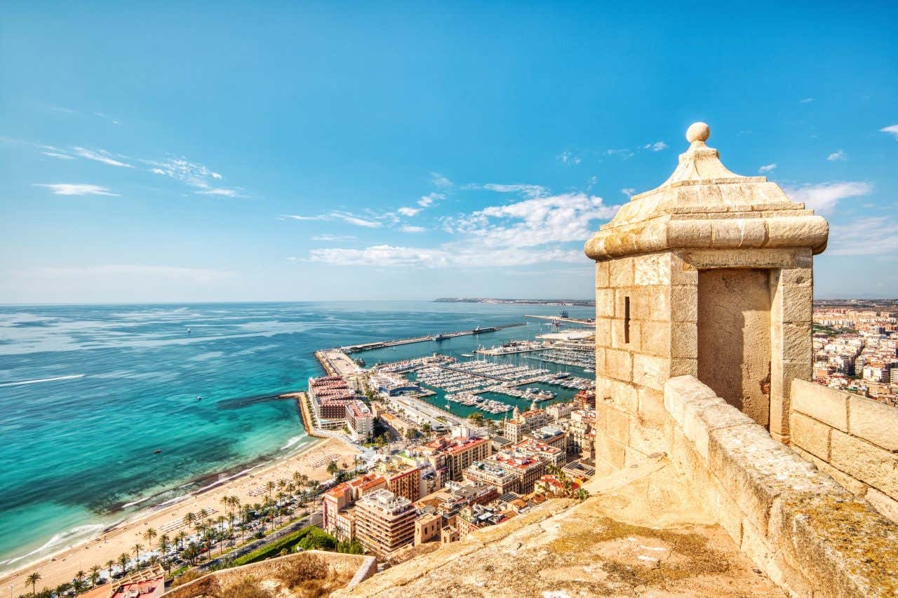 Vista panoramica dal Castello di Santa Barbara ad Alicante, con torretta di osservazione in primo piano, il porto turistico, la spiaggia del Postiguet e il Mar Mediterraneo sullo sfondo sotto un cielo sereno