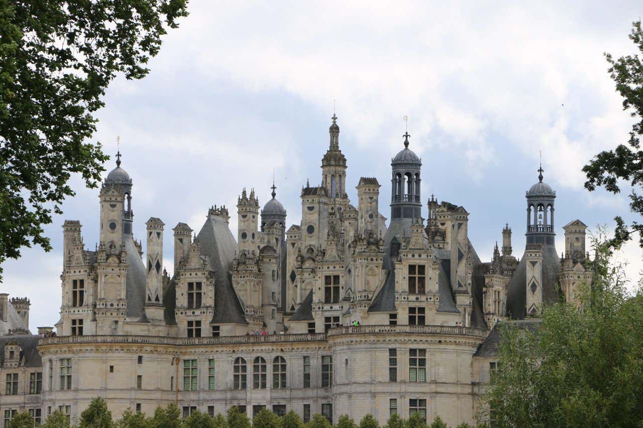 The upper half of the Château de Chambord, a large French Renaissance castle. Its roofline is characterized by many ornate spires, chimneys, and turrets, seen against a cloudy sky.