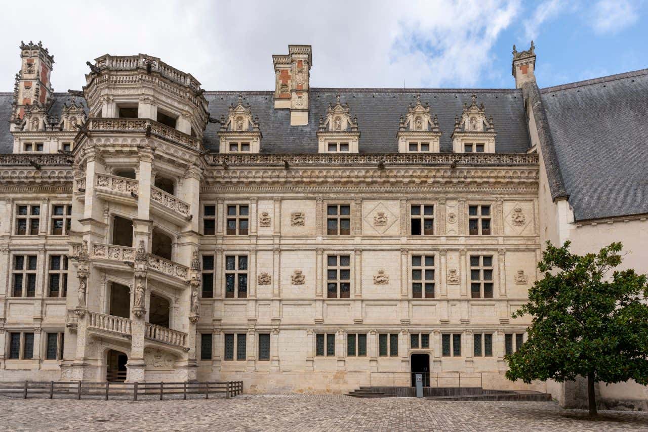 The exterior facade of a wing of the Château de Blois, featuring an ornate stone spiral staircase on the left side of the building. The castle has many windows and a grey roof, with a cobbled courtyard in the foreground.
