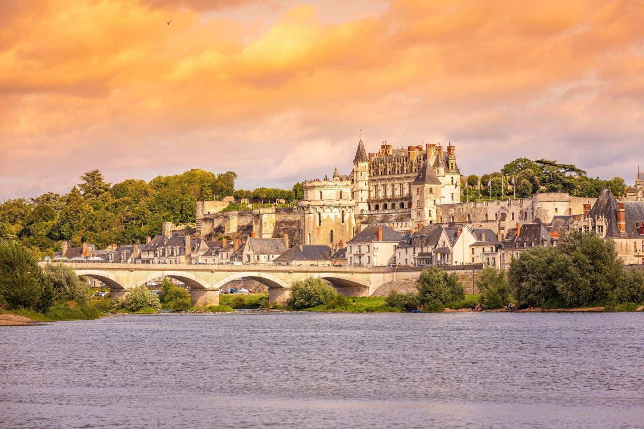 The Château d'Amboise, a large castle with a Renaissance facade, is situated on a hill overlooking the Loire River. A stone bridge crosses the river, and the sky is filled with orange and yellow clouds at sunset or sunrise.