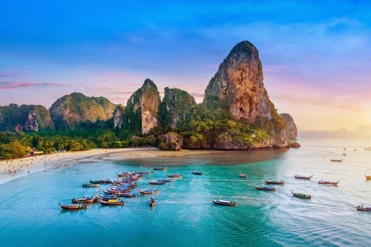 Panorama di Railay Beach a Krabi, Thailandia, con barche tradizionali dalla forma allungata, ancorate sul mare turchese, spiaggia di sabbia chiara e imponenti scogliere calcaree immerse nella vegetazione tropicale al tramonto