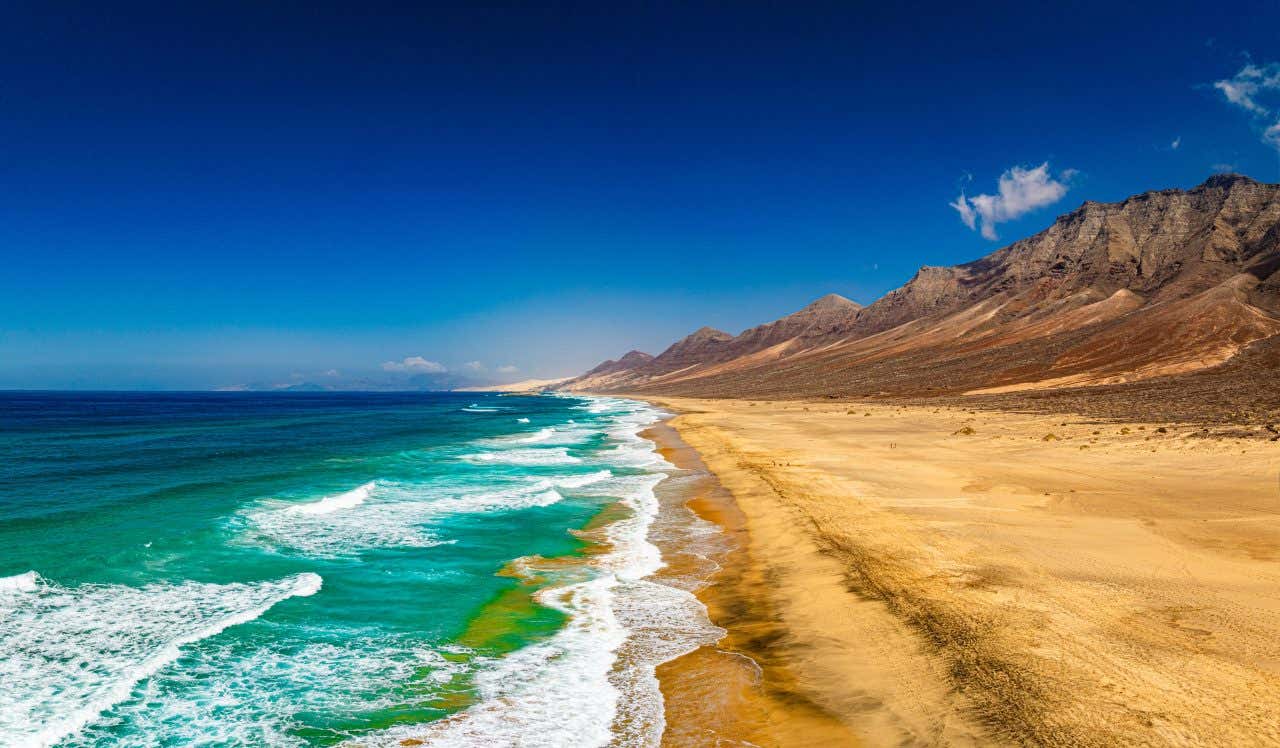 A beach with golden sand, crystal-clear sea, and rocky mountains in the distance on a sunny day.
