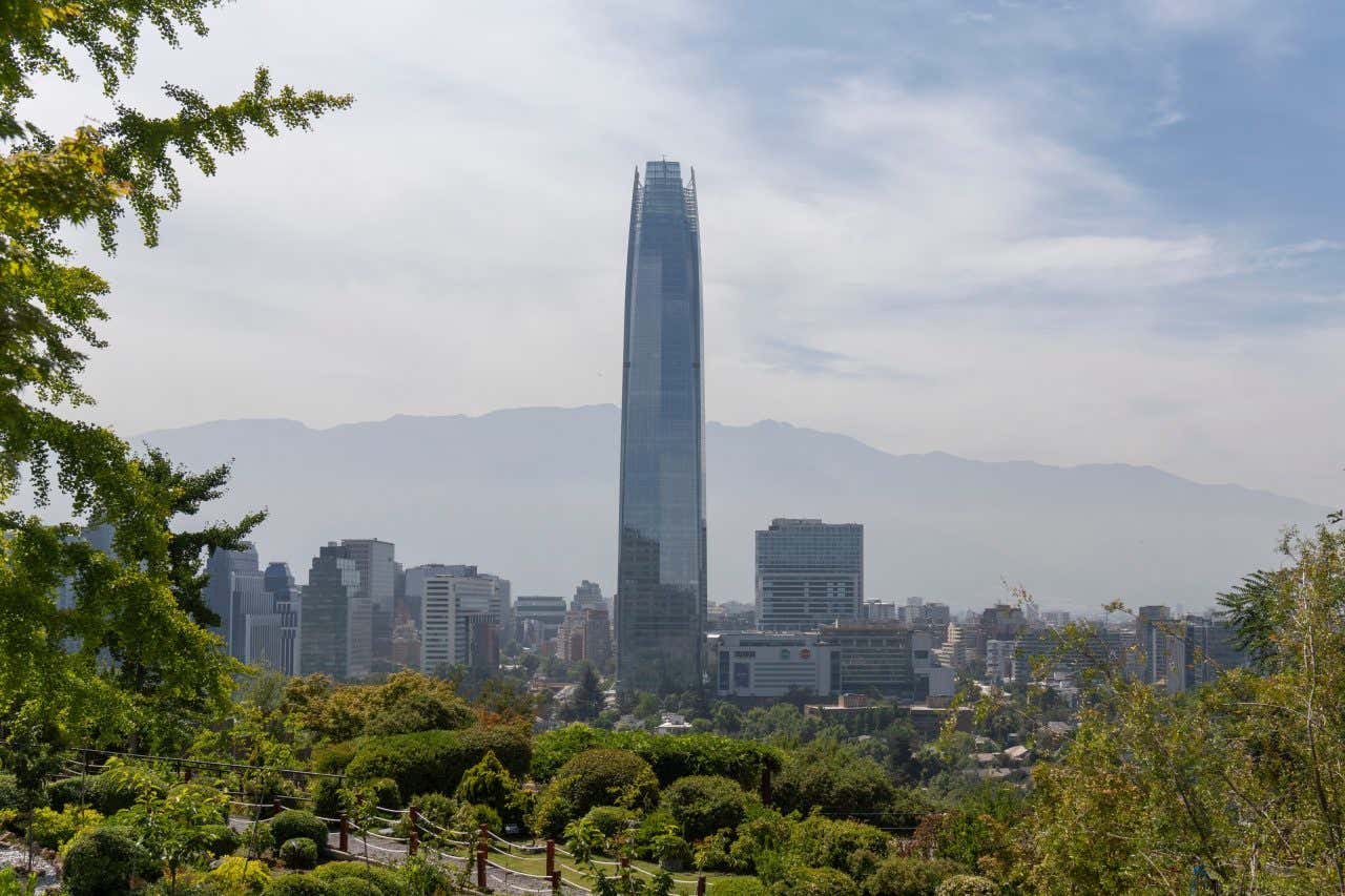 Gran Torre in Santiago, Chile as seen from afar, with a cloudy sky above and various other buildings and a mountain in the background.