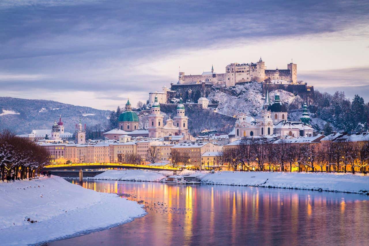 Vista di Salisburgo in dicembre, con la città avvolta dalla neve e illuminata dalle luci delle luminarie di Natale. In primo piano si vede il fiume Salzach, mentre sullo sfondo spicca la fortezza Hohensalzburg, che sovrasta la città