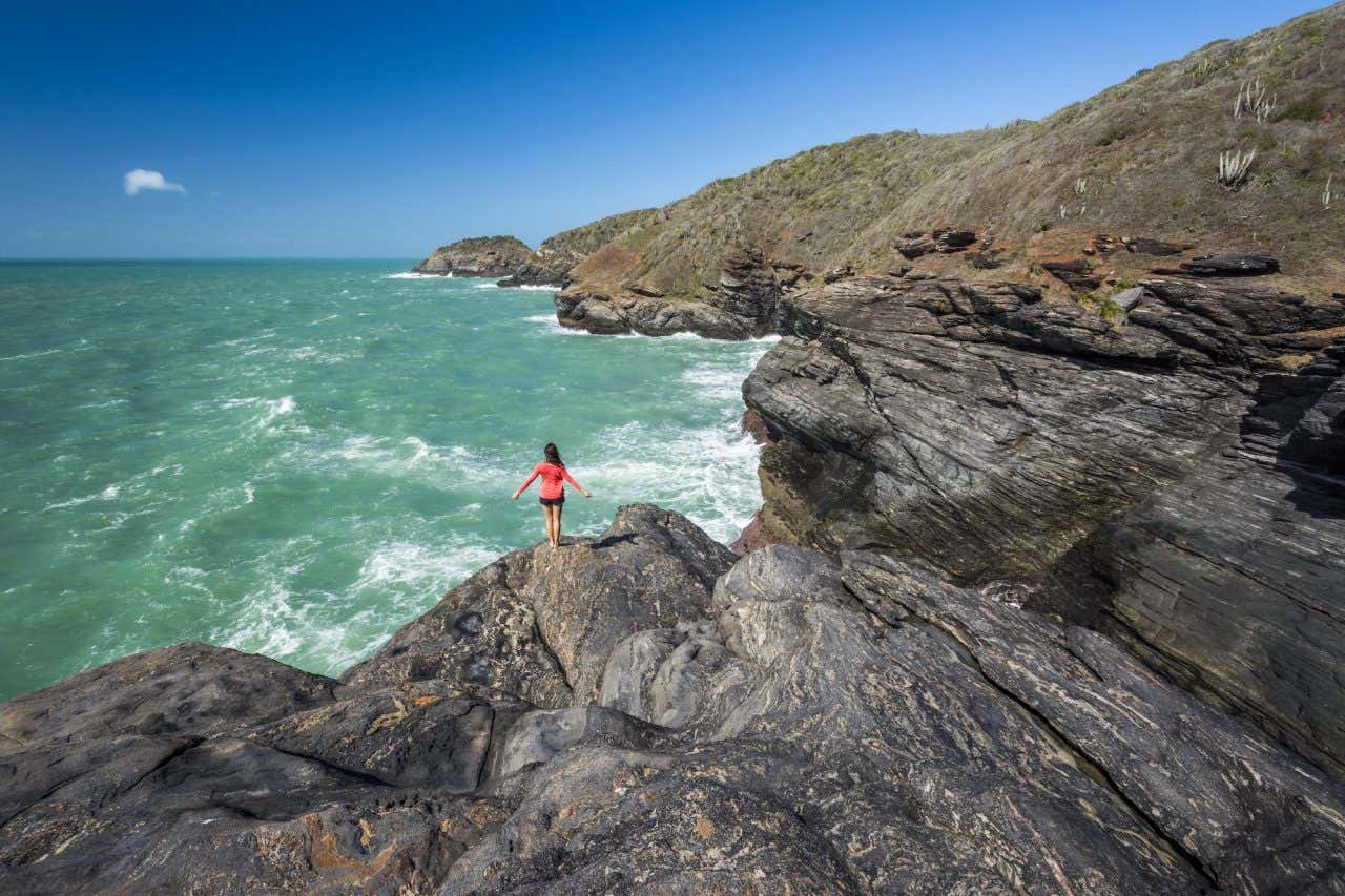 Uma mulher de costas e com os braços abertos na ponta de uma formação rochosa olhando águas verdes com o céu ensolarado